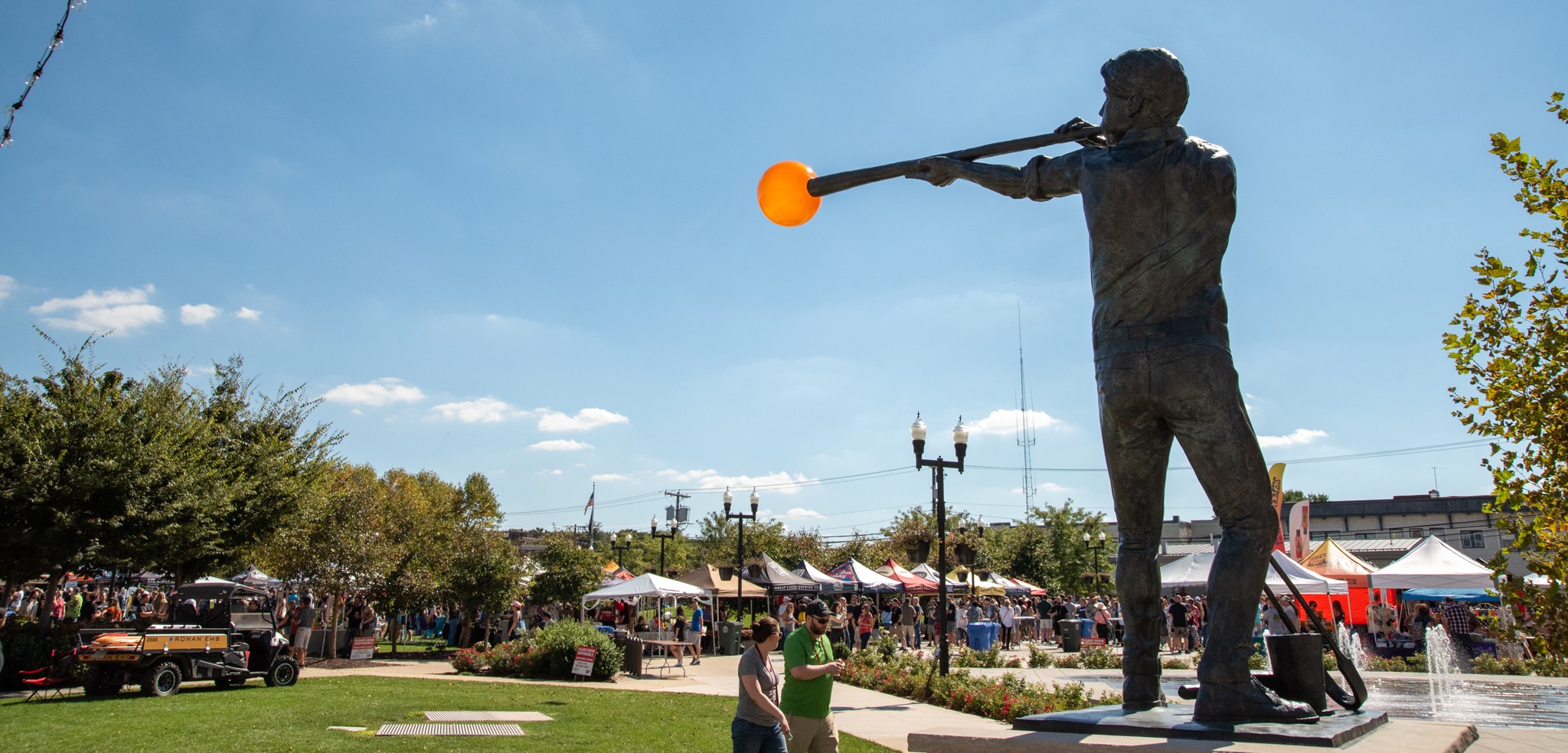 A photo of the "Glassblower" statue in Glassboro, NJ, with a community event taking place in the background